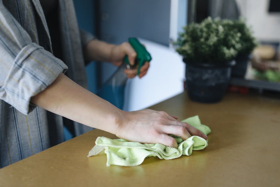 A person wearing a plaid shirt is engaged in surface cleaning within a home or office environment. Their left hand is pressing a yellow cloth to wipe a wooden table surface, which appears smooth and polished. The right hand, partly visible, is holding a green spray bottle, suggesting the application of cleaning solution. In the background, there is a potted plant with dense green foliage, and the room is illuminated by natural light, creating a bright and clean atmosphere. The scene reflects detailed domestic cleaning activities, emphasizing hygiene and surface maintenance, with emphasis on wood surface cleaning. Mayfair Carpet Cleaners' professional approach to deep cleaning and sanitisation is evident in the thoroughness of the process depicted, aligning with the Berkeley Square home-cleaning guide for Mayfair residents.