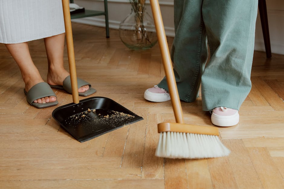 A person wearing a plaid shirt is engaged in surface cleaning within a home or office environment. Their left hand is pressing a yellow cloth to wipe a wooden table surface, which appears smooth and polished. The right hand, partly visible, is holding a green spray bottle, suggesting the application of cleaning solution. In the background, there is a potted plant with dense green foliage, and the room is illuminated by natural light, creating a bright and clean atmosphere. The scene reflects detailed domestic cleaning activities, emphasizing hygiene and surface maintenance, with emphasis on wood surface cleaning. Mayfair Carpet Cleaners' professional approach to deep cleaning and sanitisation is evident in the thoroughness of the process depicted, aligning with the Berkeley Square home-cleaning guide for Mayfair residents.
