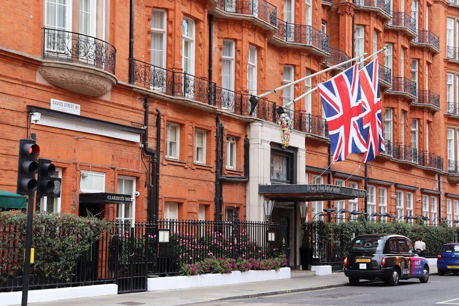 Exterior view of a red brick building with multiple floors and black wrought iron balconies, adorned with Union Jack flags hanging from the facade. The entrance features a black awning with the name 'Claridge's' visible, surrounded by a black metal fence with pink flowering bushes lining the street. A traffic light and parked cars are present in the foreground, under a clear daytime sky. This image depicts a classic London building in Grosvenor Square, emphasizing its historic architecture and vibrant city atmosphere, closely related to domestic and commercial surface cleaning needs discussed on mayfaircarpetcleaners.com.