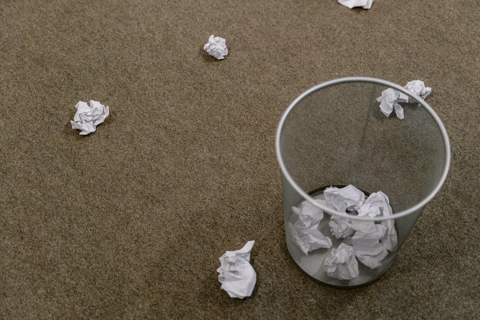 A beige carpeted floor in an office or living room setting, with several crumpled sheets of white paper scattered across the surface and inside a transparent wastebasket. The wastebasket is positioned near the center right of the image and contains additional crumpled paper. The room appears to be well-lit with natural or artificial light, highlighting the texture of the carpet and the disorganized state of the paper waste. Part of a white object or garment is visible in the top right corner. The scene reflects a typical area of surface cleaning and tidying, emphasizing the importance of waste disposal and maintaining cleanliness, as would be relevant in advice for proper bulky waste and carpet disposal for Mayfair tenants, supported by Mayfair Carpet Cleaners.