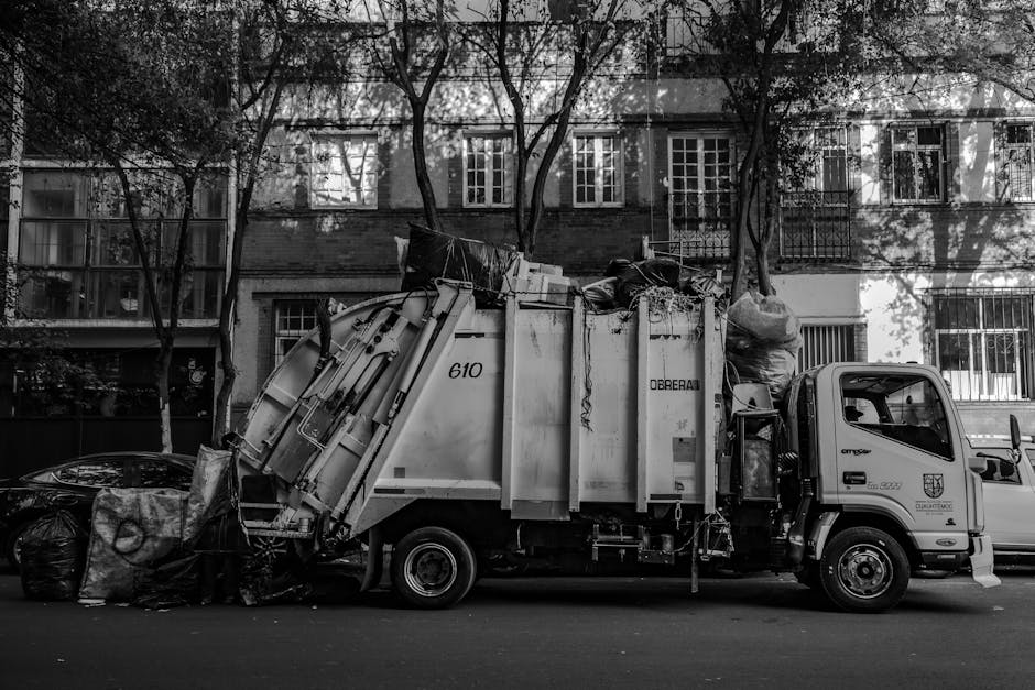 A black-and-white photograph of a waste collection truck parked on a residential street in Mayfair. The truck is loaded with bulky waste items and debris, including large bags and miscellaneous refuse, positioned against an urban backdrop of multi-storey apartment buildings with numerous windows and balconies. The street is lined with leafless trees casting shadows across the buildings and vehicle. The scene appears to be during daytime, showcasing the process of waste removal, which is relevant to advice on bulky waste disposal for Mayfair tenants, as provided by Mayfair Carpet Cleaners in their cleaning and maintenance services.