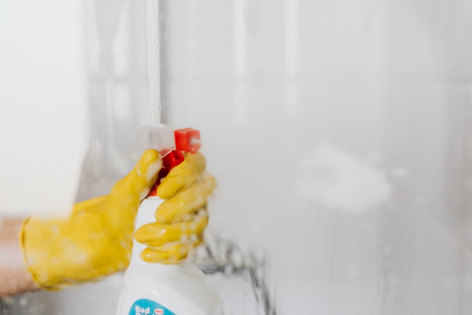 A close-up of a person's hand wearing a yellow rubber cleaning glove, holding a white spray bottle with a red nozzle, actively applying a cleaning solution to a glass shower enclosure. The glass surface is partially obscured by steam or mist, indicating recent cleaning or sanitisation. The background shows a slightly blurred bathroom setting with white tiles and soft natural light, highlighting a thorough surface cleaning process. This image aligns with the services offered by Mayfair Carpet Cleaners for emergency stain removal and domestic cleaning, emphasizing rapid and effective surface cleaning in a bathroom environment.