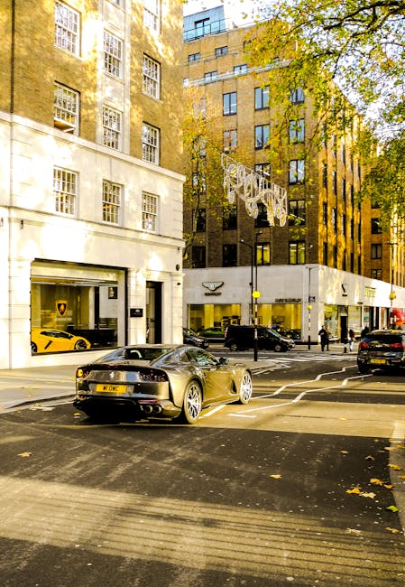 Photograph of a street scene in Mayfair West End, featuring a modern building with large windows and white facade, and a storefront with visible signage. Several luxury cars, including a black Audi an