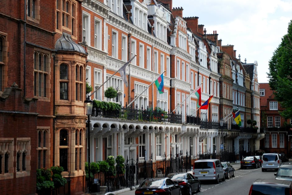 A row of historic red-brick townhouses in Mayfair, London, featuring ornate bay windows, decorative white trim, and cast-iron balconies with potted plants. Several flags are mounted on the balconies, and the street below has parked cars along the curb under the soft daylight. The buildings are well-maintained with clean facades, reflecting a classic British architectural style. The image highlights residential exterior surfaces, with attention to the neatness and aesthetic details typical of upscale properties, aligning with Mayfair Carpet Cleaners' focus on surface cleaning and property maintenance.