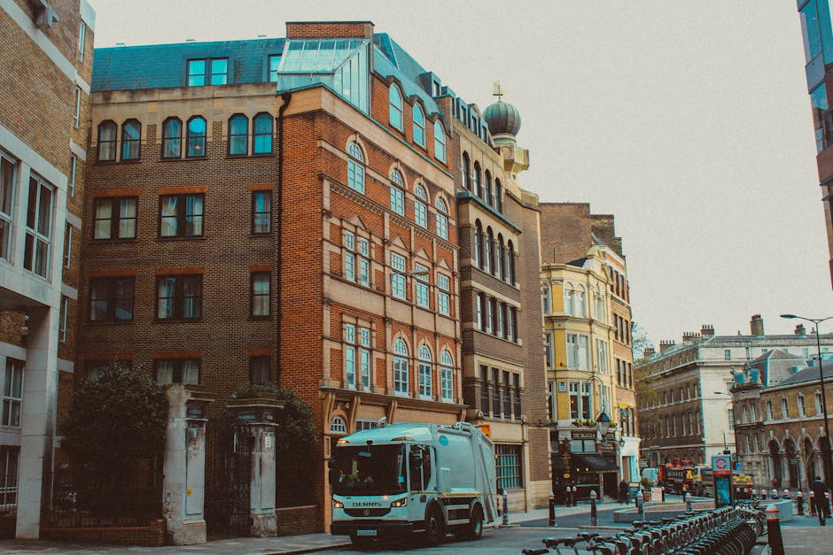 A street scene in Westminster with historic multi-story buildings featuring brick and ornate facades, some with arched windows and decorative elements. The pavement is clean and wet, possibly from recent surface cleaning, and a modern street cleaning vehicle is parked on the side, indicating ongoing urban hygiene maintenance. The lighting suggests a cloudy day, with soft natural light illuminating the area. This scene highlights the importance of surface cleaning and deep cleaning in maintaining the visual appeal and hygiene standards of Westminster properties, in line with Westminster Council rules on cleaning waste for Mayfair homes. Mayfair Carpet Cleaners specialises in professional domestic and commercial cleaning, ensuring thorough sanitisation of surfaces such as stone, brick, and glass in historical and modern buildings.