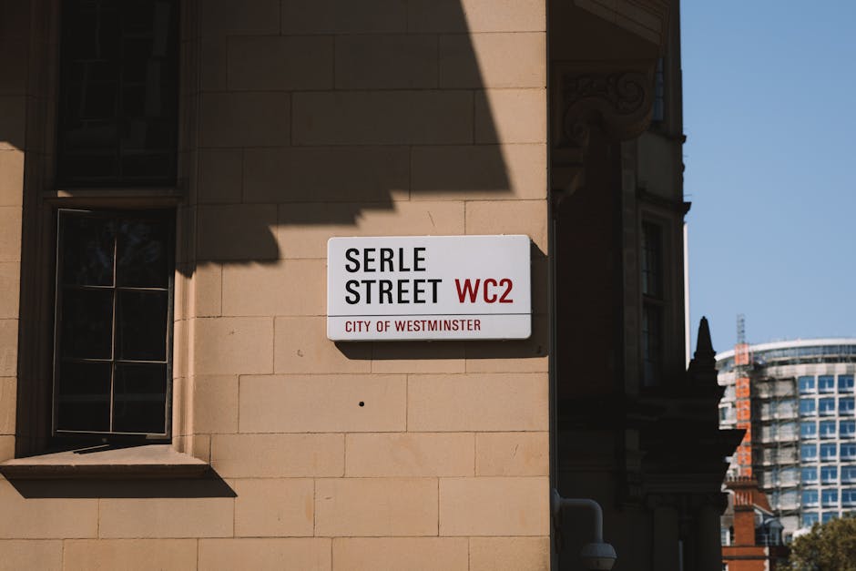 A close-up view of a rectangular street sign mounted on a beige stone building wall in Westminster. The sign reads 'SERLE STREET WC2 CITY OF WESTMINSTER' with black and red lettering on a white background. The building's window with dark framing is visible to the left, and a shadow from an overhanging structure partially covers the top part of the sign. In the background, modern high-rise buildings and a clear blue sky are visible, indicating an urban environment with bright natural lighting.